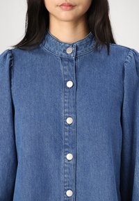 Denim shirt in medium blue with a high collar, silver snap buttons down the front, and gathered puff sleeves at the shoulders.