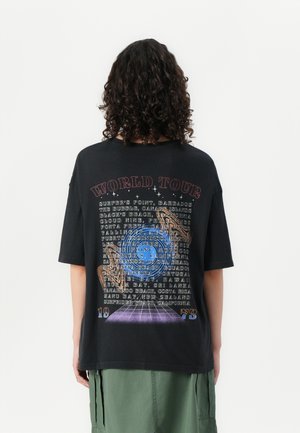 Camiseta negra de gran tamaño con un gráfico colorido de "World Tour" que incluye varias ubicaciones, diseñada con un ajuste relajado y mangas cortas.