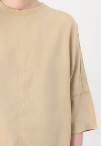 Beige, short-sleeve shirt made of soft fabric with a round neckline and visible seam detail. The fit is relaxed with dropped shoulder seams.