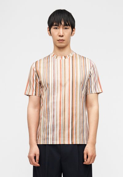 Man som bär en kortärmad T-shirt med vertikala flerfärgade ränder och svarta byxor, stående mot en enfärgad vit bakgrund.