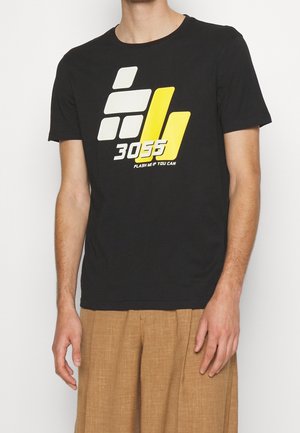 Homme portant un t-shirt noir avec des formes géométriques blanches et jaunes, le numéro 3055, et le texte "FLASH ME IF YOU CAN", assorti à un pantalon beige.