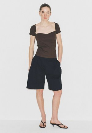 Kvinde står mod hvid baggrund iført en brun tætsiddende top med korte ærmer, sorte knælange shorts og sorte sandaler med hæl.