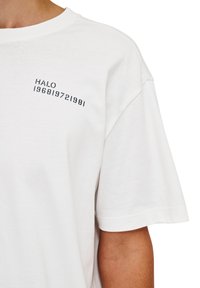 Biała bawełniana koszulka z okrągłym dekoltem i krótkimi rękawami, ozdobiona czarnym napisem "HALO 196819721981" na klatce piersiowej.
