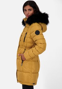 Vrouw in een mosterdgeel gewatteerd jack met een zwart bontkraag, handen in de zakken, kijkend naar de camera op een witte achtergrond.