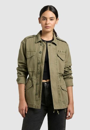 Junge Frau mit dunklen Haaren im Pferdeschwanz, trägt eine olivgrüne, geknöpfte Jacke über einem schwarzen Crop-Top und dunklen Jeans, steht vor einem schlichten Hintergrund.