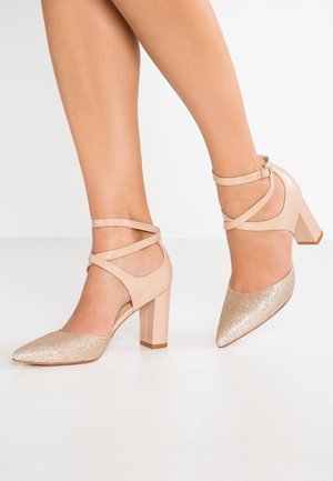 Zapatos de tacón alto beige con correas en el tobillo, con punta dorada brillante y tacón grueso. Textura de cuero suave en todo el zapato.