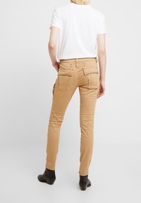 Beige cargo-housut, joissa on kapea leikkaus, useita taskuja ja koristeellista metalliosaa. Yhdistetty valkoisen t-paidan ja mustien nilkkabootsien kanssa.