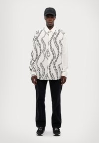 Camisa blanca de manga larga con patrón de cadenas negras, combinada con pantalones negros y zapatillas negras, rematada con una gorra negra con logo.