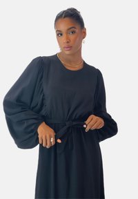 Vestido negro con mangas abullonadas, escote redondo y cinturón anudado en la cintura. Tela suave con una textura fluida. Detalles de diseño minimalistas.