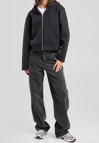 Černá krátká mikina s kapucí na zip vpředu, s volným oversized střihem a předními kapsami. Kombinováno s volnými tmavě šedými džíny a šedými teniskami.