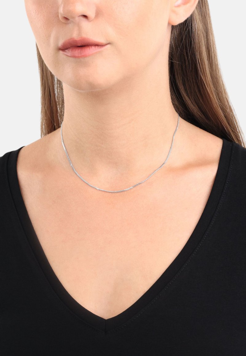 Silverfärgad kedjehalsband med en delikat, slät textur, som har en enkel design. Bärs av en person med en svart topp med V-ringning.