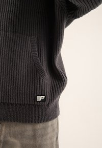 Dunkelgrauer gerippter Pullover mit einer Tasche, ausgestattet mit einem schwarzen Etikett und einem weißen Logo. Weiche Textur, lässiges Design und enge Passform.