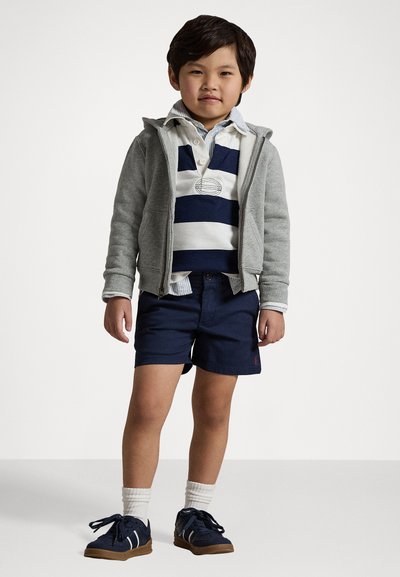 Grau zip-up Hoodie, weiß und marineblau gestreiftes Polo, marineblaue Shorts, weiße Sneaker-Socken und marineblaue Turnschuhe mit braunen Sohlen. Einfaches und lässiges Outfit.