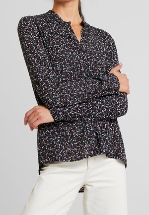 Woman wearing a long-sleeve black shirt with small red, white, and blue floral pattern and off-white pants, arms crossed.
