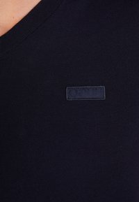 Maglione a V in maglia di colore navy, caratterizzato da un patch logo testurizzato. Il tessuto è liscio, con una trama fine e fitta e un colletto costolato.