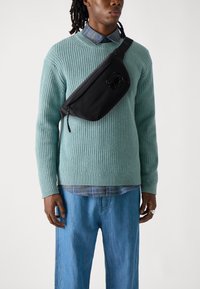 Black fabric sling bag with a glossy logo, worn over a light green ribbed knit sweater and layered with a blue checkered shirt beneath.