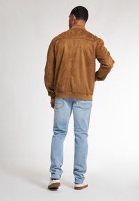 Giacca bomber in suede marrone con colletto a coste e dettagli sulle cuciture posteriori, indossata sopra jeans azzurri chiari e sneakers bianche, vista da dietro.