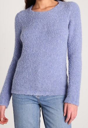 Femme portant un pull à manches longues texturé violet clair associé à un jean en denim bleu clair, montrée du cou aux hanches.