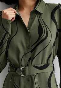 Camisa verde de botones con remolinos abstractos en negro, con cintura ceñida y mangas largas. Tela suave con una textura lisa.