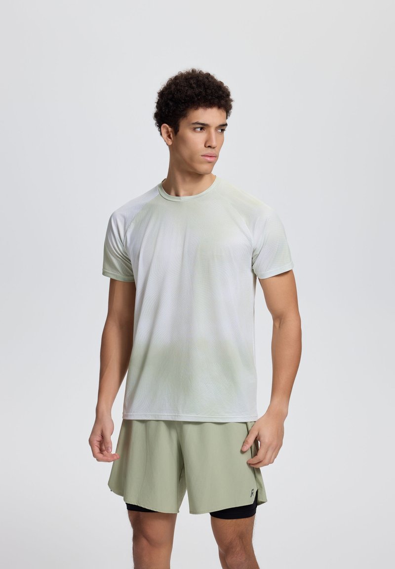 Junger Mann steht, trägt hellgrüne Sportshorts über schwarzen Leggings und ein hellgraues kurzärmeliges Sportshirt, schaut zur Seite.