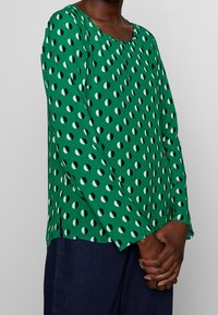 Grünes Langarmshirt mit Polka-Dot-Muster aus abwechselnd weißen und grünen Hälften. Rundhalsausschnitt und seitliche Schlitze am Saum.