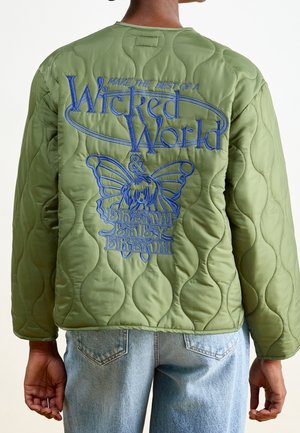 Person iført grøn quiltet jakke med blå broderet tekst "Make the best of a Wicked World" og sommerfugldesign på ryggen.