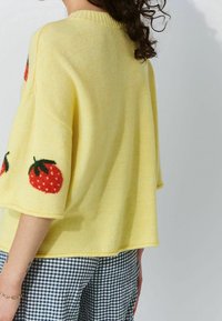 Femme portant un pull jaune ample avec des motifs de fraises rouges sur les manches et un pantalon à carreaux noir et blanc, vue de dos.