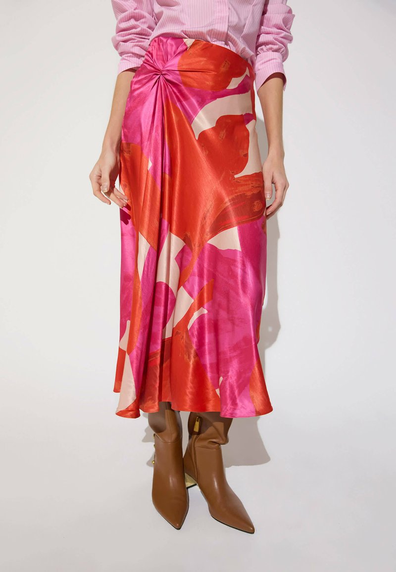 Woman wearing a pink and red abstract patterned satin skirt with a twisted knot detail, paired with tan pointed-toe ankle boots and a pink shirt.
