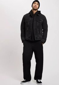 Czarna kurtka dżinsowa z dwoma kieszeniami na piersi, nałożona na czarny hoodie. Połączona z czarnymi spodniami o szerokich nogawkach i czarnymi trampkami.