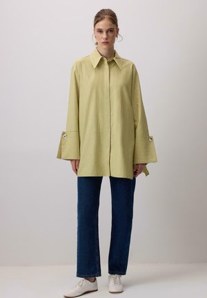 Mladá žena stojaca, oblečená v voľnej žltej pruhovanej košeli so širokými manžetami, tmavomodrých džínsoch a bielych teniskách na jednoduchom pozadí.