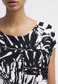 Camicia a righe in bianco e nero con scollo a barca, maniche corte e taschino frontale con un design floreale astratto.