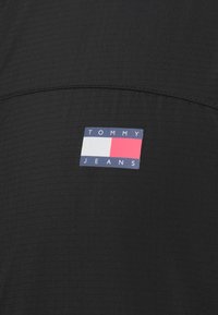 Μαύρο ύφασμα με υφή, με ένα ορθογώνιο λογότυπο σε ναυτικό μπλε, κόκκινο και άσπρο που γράφει "TOMMY JEANS".