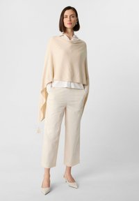 Beige gebreide poncho met een losse vorm, gedragen over een witte blouse met knopen, gecombineerd met crèmekleurige broeken en beige puntige hakken. Heeft franjes.