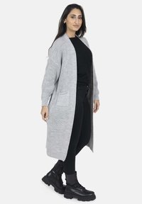 Cardigan lungo grigio a maglia con due tasche frontali e design aperto, abbinato a jeans skinny neri e stivaletti.
