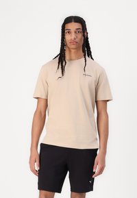 Beige Baumwoll-T-Shirt mit Rundhalsausschnitt und kurzen Ärmeln, mit einem kleinen Columbia-Logo auf der linken Brust. Getragen mit schwarzen Shorts.