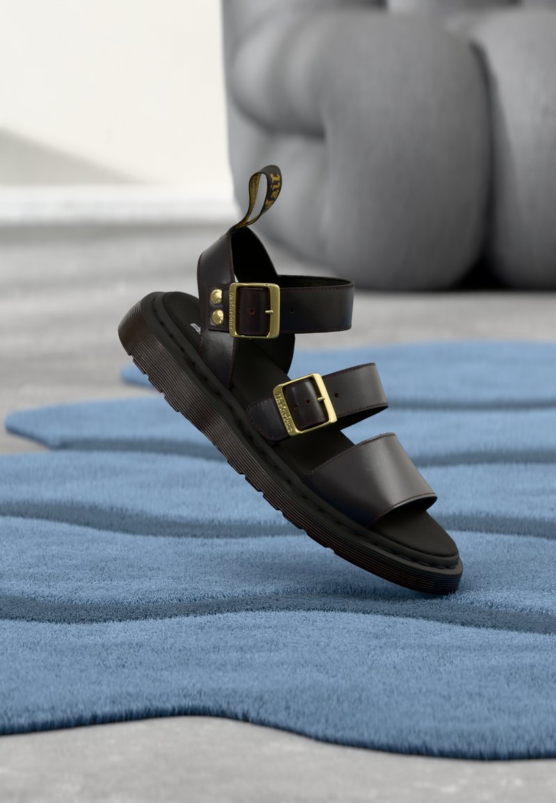 Σκούρο καφέ δερμάτινο σανδάλι με δύο χρυσές αγκράφες και χοντρή σόλα, τοποθετημένο πάνω σε υφασμένο μπλε χαλί και γκρι φόντο.