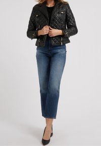 Veste de moto en cuir matelassé noir avec quincaillerie dorée, poches zippées et design ajusté, associée à un jean bleu taille mi-haute.