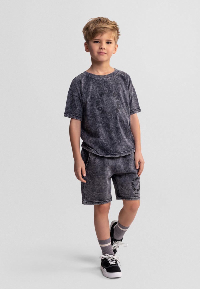 Niño de pie con una pierna cruzada, vistiendo una camiseta negra desgastada y pantalones cortos a juego, zapatillas negras y calcetines grises.