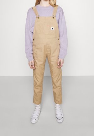 Personne portant une salopette beige, un sweat-shirt violet clair et des baskets montantes blanches, debout devant un fond uni.
