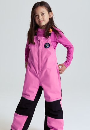 Ung pige står med hænderne på hoften iført lyserøde og sorte vandtætte overalls og en langærmet lyserød trøje mod en ensartet baggrund.