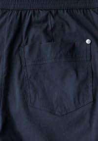 Tissu bleu marine foncé avec une texture à fines côtes. Présente une poche arrière avec un rabat arrondi et un accent de bouton-pression. Ceinture élastique.