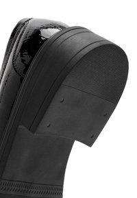 Schwarze Lacklederschuhe mit einer klobigen Gummisohle. Die Ferse hat eine strukturierte Oberfläche und ist rund geformt mit vertikalen Rillen.