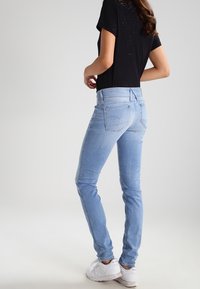Světlé modré džíny s úzkým střihem, které mají klasický pětikapsový design, jemné vyblednutí a střední pas. Nosič má na sobě černý top.