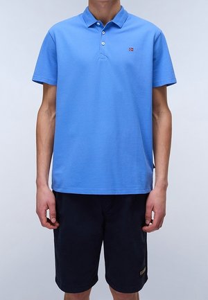 Ein Mann trägt ein hellblaues kurzärmeliges Poloshirt und dunkelblaue knielange Shorts und steht vor einem einfarbigen Hintergrund.