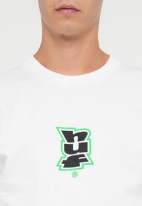 Hvit bomulls t-skjorte med en svart og grønn "huf" logo i midten. Rund hals og korte ermer, med en glatt tekstur.