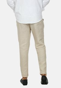 Pantaloni di lino beige con un design a gamba dritta, polsini arrotolati e una texture liscia. Indossati con una camicia bianca a maniche lunghe e calzature nere.