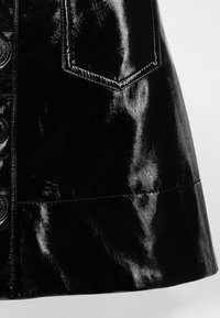 Mustast lakknahast seelik läikiva viimistlusega, millel on ees taskud, nööpidega sulgemine ja struktureeritud siluett.