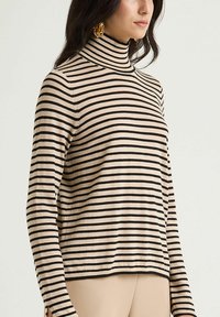 Frau trägt einen beige-schwarz horizontal gestreiften Rollkragenpullover, kombiniert mit beigen Hosen und goldenen Creolen, seitlich stehend.