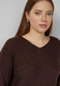 Brązowy sweter dziany z dekoltem w serek, o fakturowanym materiale i luźnym kroju. Modelka nosi duże srebrne kolczyki koła.