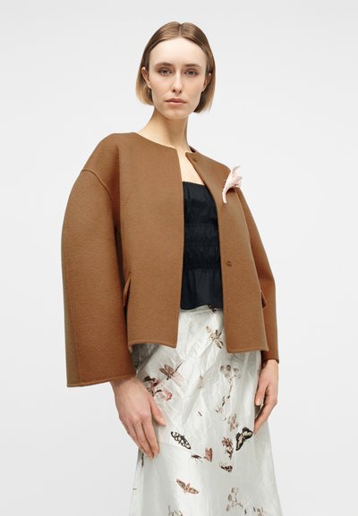 Femme portant une veste marron oversize avec une épingle fleur rose, un haut noir et une jupe blanche imprimée de papillons et de fleurs, sur un fond uni.